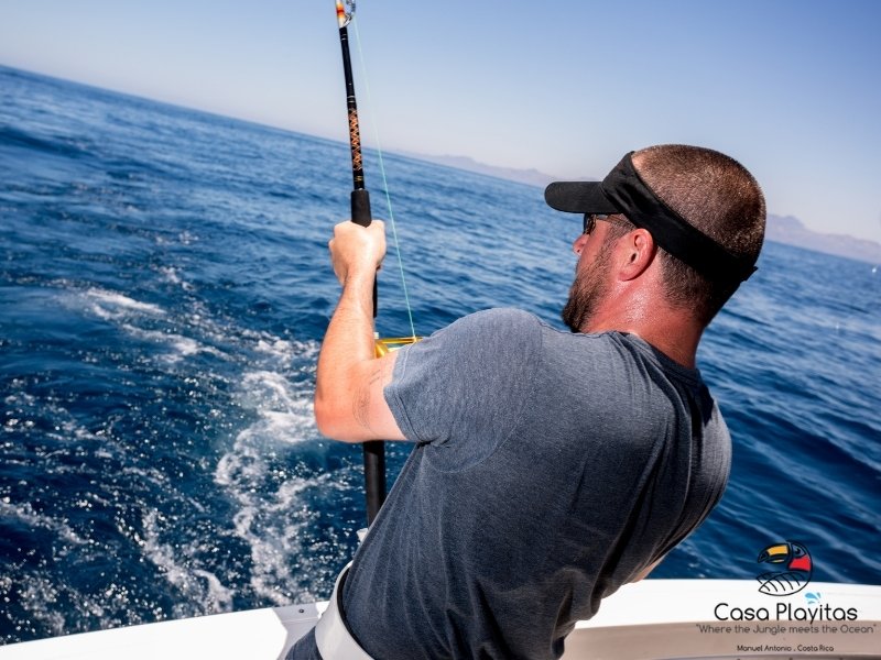 Man wearing a gray shirt and visor reels in a fishing rod from the side of a boat on a deep blue ocean under a clear sky.