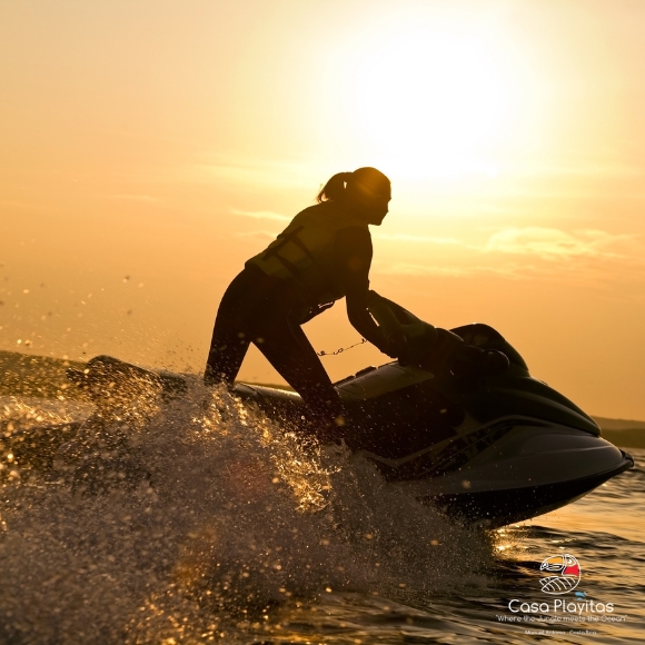 Silhouette of two people riding a jet ski at sunset, spray flying off the waves.